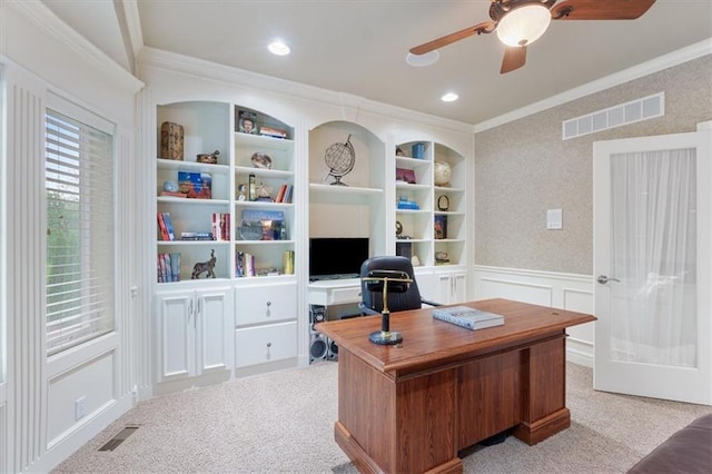 home office featuring a ceiling fan, ornamental molding, carpet floors, a decorative wall, and wainscoting