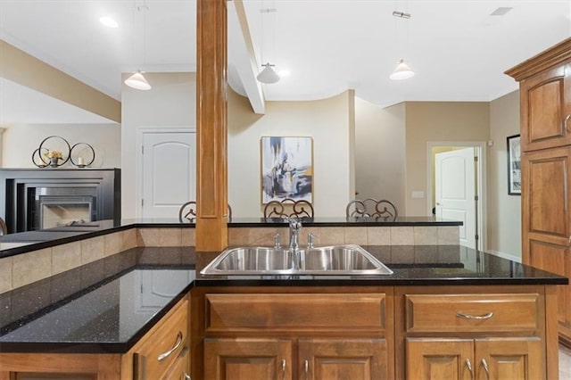 kitchen with a sink, brown cabinetry, hanging light fixtures, dark stone counters, and a peninsula