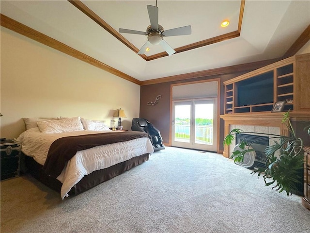 carpeted bedroom with access to exterior, a tiled fireplace, a ceiling fan, a raised ceiling, and ornamental molding