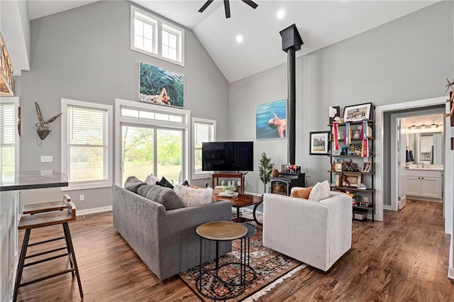 living area with dark wood-style flooring, a wood stove, high vaulted ceiling, a ceiling fan, and baseboards
