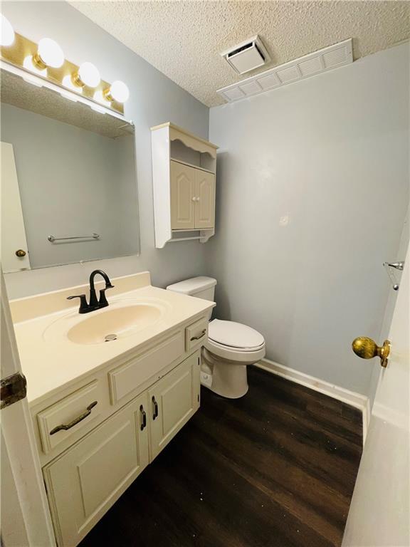 half bathroom with vanity, toilet, a textured ceiling, wood finished floors, and visible vents