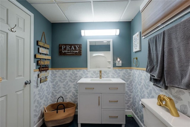 half bathroom featuring a paneled ceiling, wainscoting, vanity, toilet, and wallpapered walls
