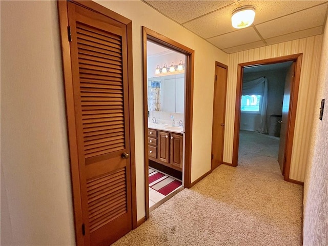 hallway featuring baseboards, a sink, light carpet, and a paneled ceiling