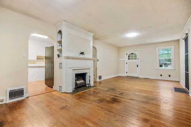 unfurnished living room with light wood-style floors, a brick fireplace, arched walkways, baseboards, and visible vents