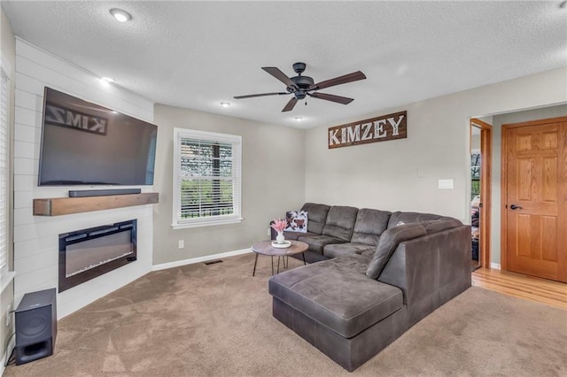 carpeted living room featuring a ceiling fan, a textured ceiling, visible vents, baseboards, and a glass covered fireplace