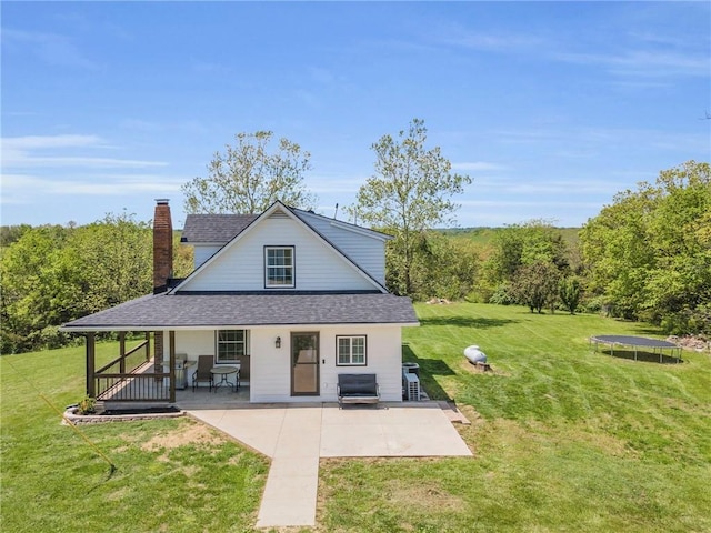 back of house with a porch, roof with shingles, a trampoline, a chimney, and a lawn