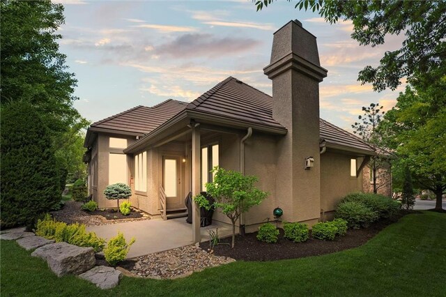 back of property at dusk featuring a chimney, a tiled roof, stucco siding, and a lawn