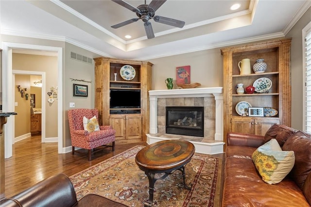 living area with a tray ceiling, wood finished floors, a fireplace, ceiling fan, and ornamental molding