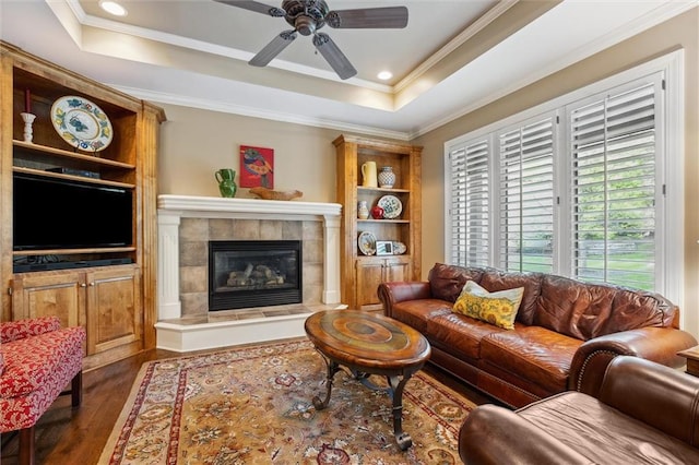 living room featuring wood finished floors, ornamental molding, a ceiling fan, a fireplace, and a tray ceiling
