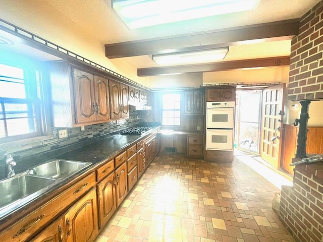 kitchen featuring under cabinet range hood, a sink, decorative backsplash, dark countertops, and double oven