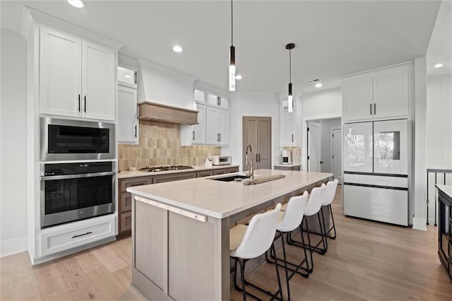 kitchen featuring stainless steel appliances, a sink, custom range hood, decorative backsplash, and a center island with sink