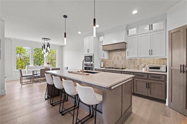 kitchen with appliances with stainless steel finishes, custom range hood, a sink, an island with sink, and backsplash