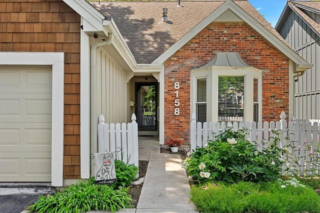 view of exterior entry with an attached garage, fence, roof with shingles, and brick siding