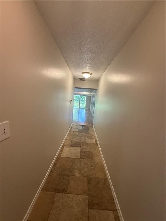 hallway with a textured ceiling, visible vents, baseboards, and stone finish floor