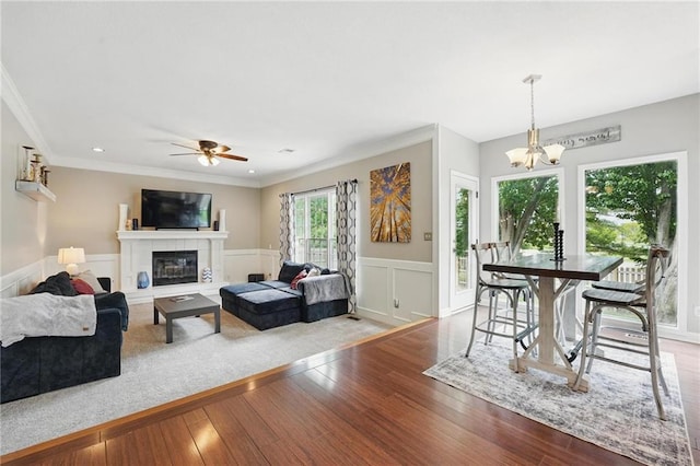 living room with wood finished floors, ceiling fan, a chandelier, a tiled fireplace, and a wainscoted wall