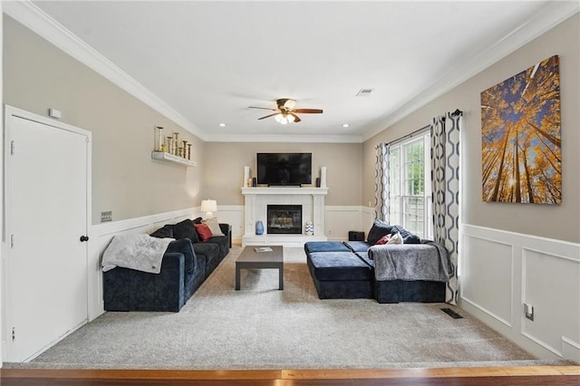 carpeted living room with a ceiling fan, ornamental molding, a glass covered fireplace, a wainscoted wall, and recessed lighting