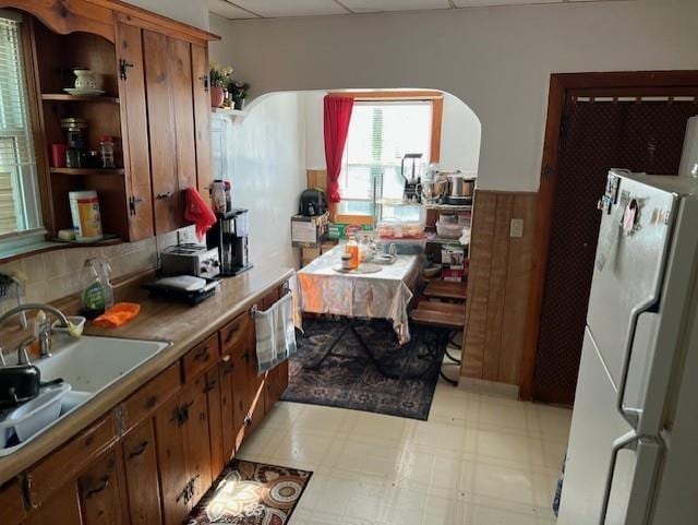 kitchen featuring a sink, brown cabinetry, freestanding refrigerator, light floors, and arched walkways