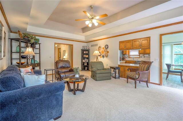 living area featuring a tray ceiling, ceiling fan, light carpet, ornamental molding, and baseboards