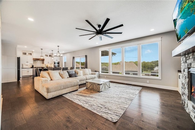 living area featuring a stone fireplace, a ceiling fan, recessed lighting, dark wood-type flooring, and baseboards