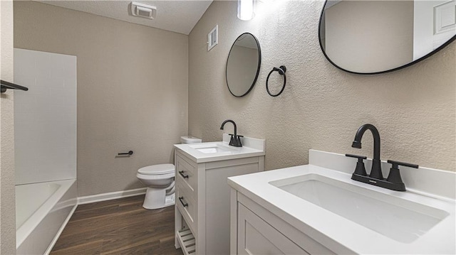 full bath with wood finished floors, toilet, two vanities, a textured wall, and baseboards