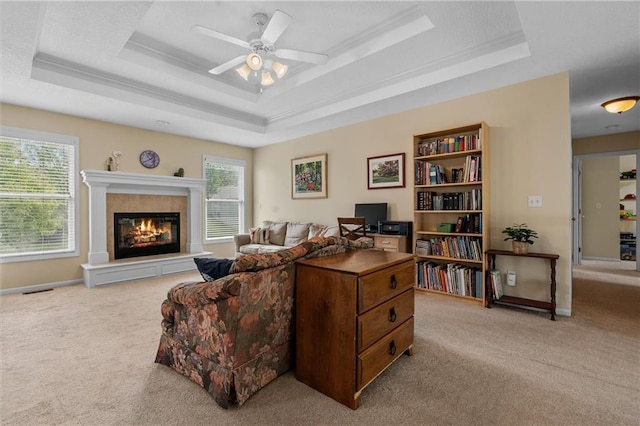 carpeted living area featuring a tray ceiling, a glass covered fireplace, ceiling fan, ornamental molding, and baseboards