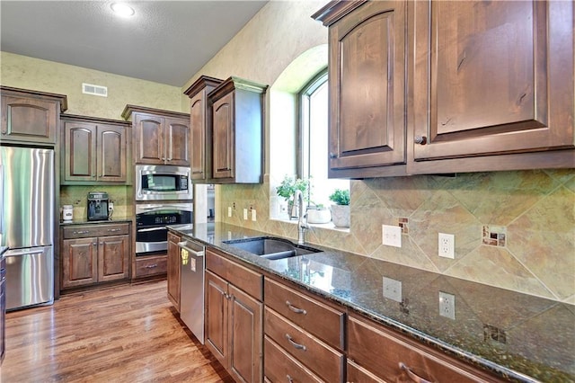 kitchen with appliances with stainless steel finishes, a sink, light wood-type flooring, dark stone counters, and backsplash