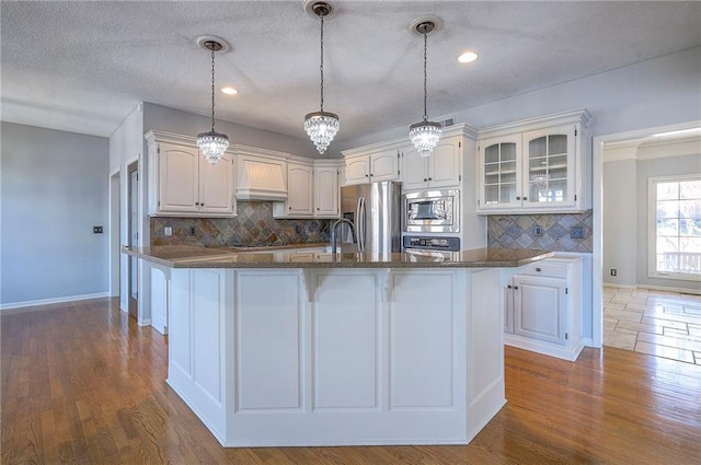 kitchen with custom exhaust hood, stainless steel appliances, glass insert cabinets, white cabinets, and tasteful backsplash
