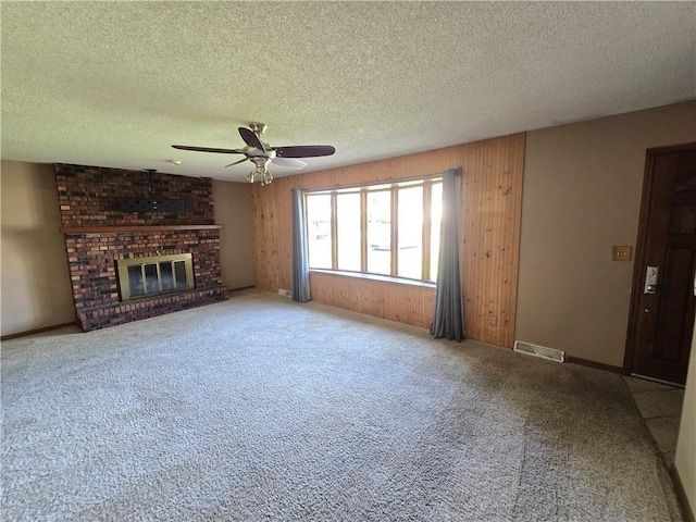 unfurnished living room featuring ceiling fan, a textured ceiling, a brick fireplace, wooden walls, and carpet flooring