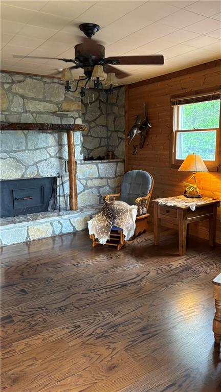 living area with ceiling fan, hardwood / wood-style floors, wood walls, and a stone fireplace
