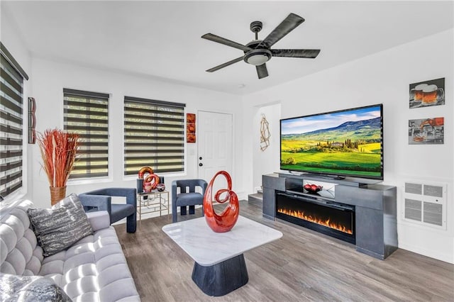living room with wood finished floors, a warm lit fireplace, and ceiling fan