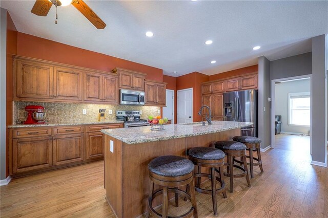 1906 Blackbird Cir, Kearney, MO 64060 - kitchen featuring stainless steel appliances, brown cabinetry, light wood-type flooring, a center island with sink, and decorative backsplash