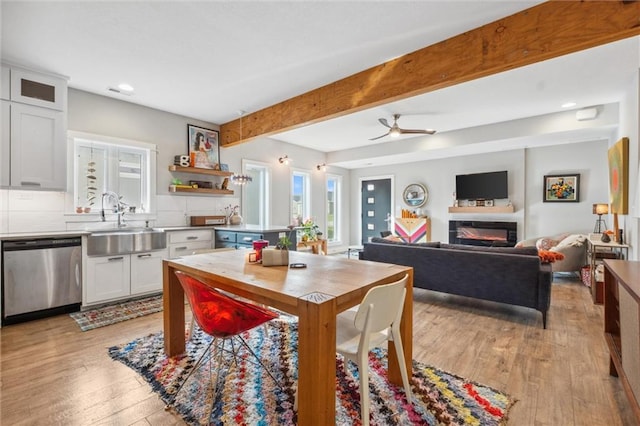 dining room featuring healthy amount of natural light, light wood finished floors, a ceiling fan, a glass covered fireplace, and beamed ceiling