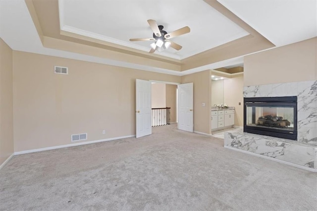 unfurnished living room with a tray ceiling, a ceiling fan, a high end fireplace, light carpet, and crown molding