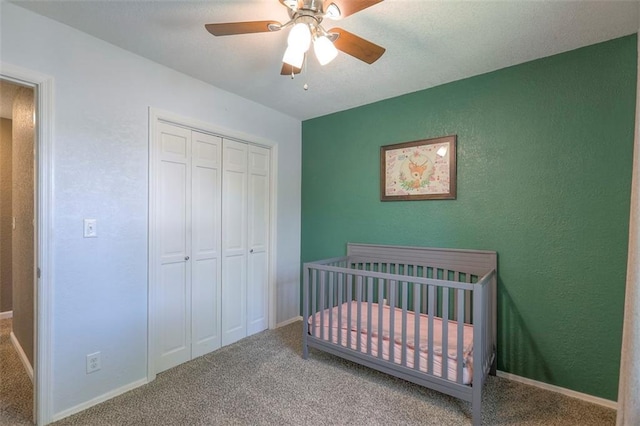 carpeted bedroom featuring a closet, a crib, a ceiling fan, baseboards, and a textured wall