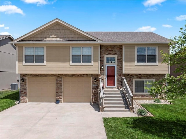 raised ranch featuring concrete driveway, a garage, central AC unit, a front yard, and roof with shingles