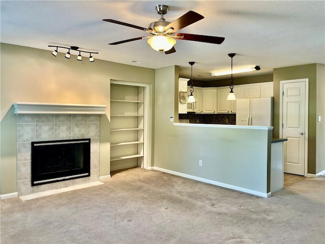 unfurnished living room with ceiling fan, a tiled fireplace, a textured ceiling, baseboards, and light colored carpet