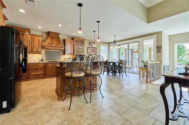 kitchen featuring freestanding refrigerator, premium range hood, brown cabinetry, a kitchen bar, and tasteful backsplash