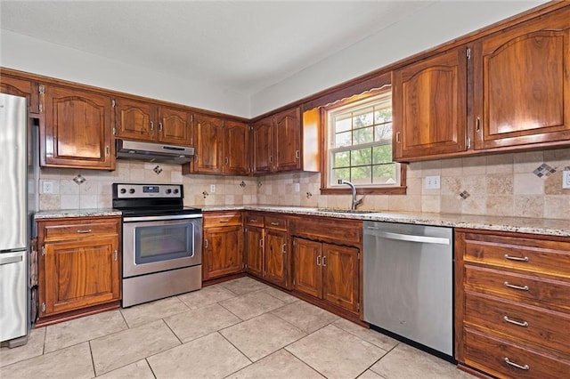 kitchen featuring stainless steel appliances, under cabinet range hood, a sink, light stone countertops, and backsplash