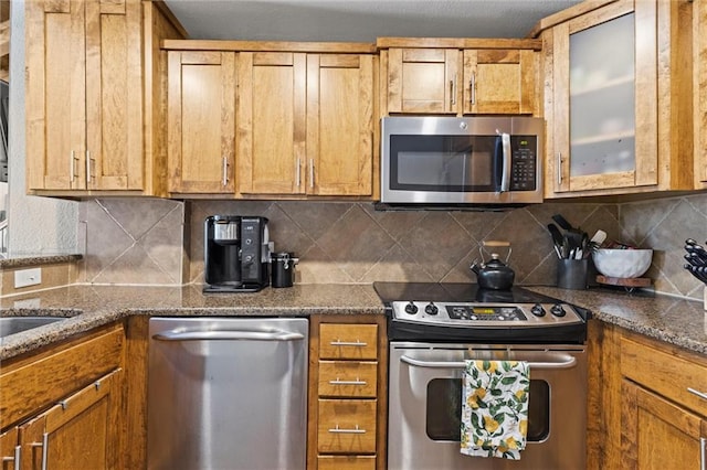 kitchen with stainless steel appliances, tasteful backsplash, glass insert cabinets, brown cabinetry, and dark stone counters