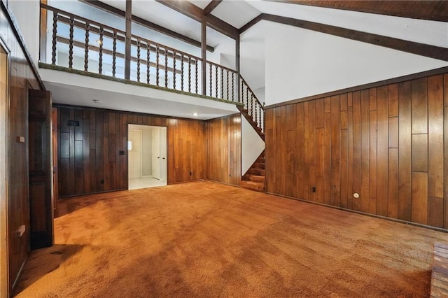 unfurnished living room with high vaulted ceiling, wooden walls, stairway, carpet flooring, and beamed ceiling