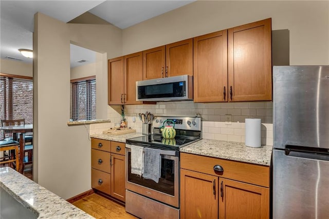 kitchen featuring appliances with stainless steel finishes, brown cabinets, backsplash, light wood finished floors, and light stone countertops