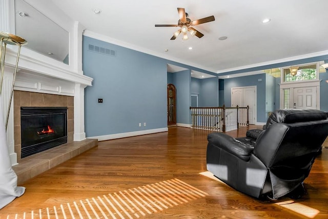 living room featuring wood finished floors, ceiling fan, a tiled fireplace, baseboards, and crown molding