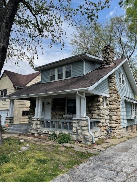view of front of house with covered porch, stone siding, a chimney, and a shingled roof
