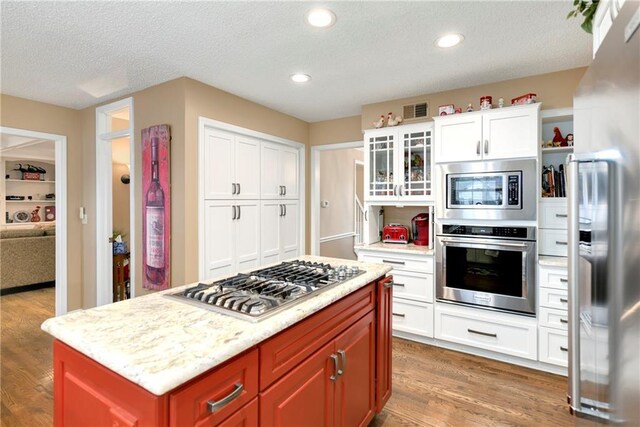 10500 Reeder St, Overland Park, KS 66214 - kitchen featuring appliances with stainless steel finishes, wood finished floors, a textured ceiling, glass insert cabinets, and white cabinetry