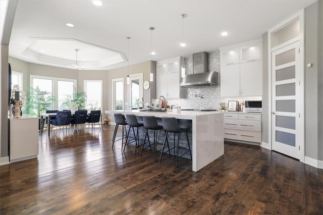 8605 NE Shl Creek Pkwy, Kansas City, MO 64157 - kitchen with wall chimney range hood, a tray ceiling, white cabinets, an island with sink, and recessed lighting