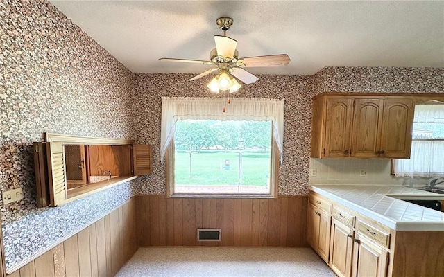 917 Country Club Dr, Butler, MO 64730 - kitchen with tile countertops, a wainscoted wall, ceiling fan, wooden walls, and a textured ceiling