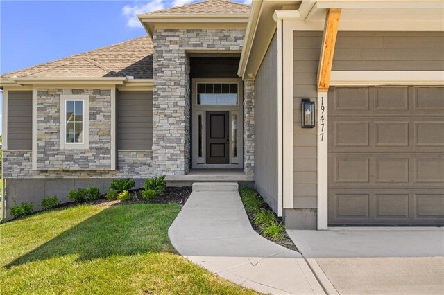 19477 W 114th Ter, Olathe, KS 66061 - view of exterior entry featuring a garage, a yard, stone siding, and roof with shingles