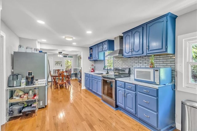 8303 Wabash Ave, Kansas City, MO 64132 - kitchen with appliances with stainless steel finishes, wall chimney exhaust hood, blue cabinetry, a ceiling fan, and light wood finished floors