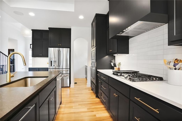 1245 NE Goshen Dr, Lees Summit, MO 64064 - kitchen featuring wall chimney range hood, stainless steel appliances, backsplash, light wood-type flooring, and dark cabinetry