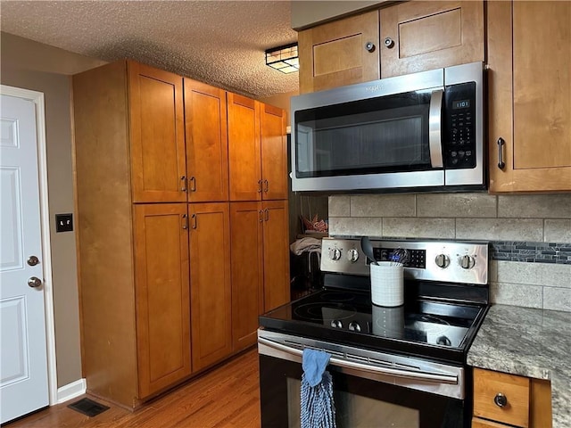 26609 S Red Bud Rd, Freeman, MO 64746 - kitchen featuring appliances with stainless steel finishes, brown cabinetry, wood finished floors, decorative backsplash, and a textured ceiling